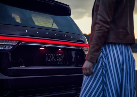 A person is shown near the rear of a 2024 Lincoln Aviator® SUV as the Lincoln Embrace illuminates the rear lights | Wallace Lincoln in Fort Pierce FL