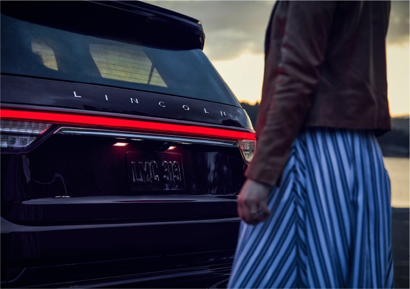 A person is shown near the rear of a 2023 Lincoln Aviator® SUV as the Lincoln Embrace illuminates the rear lights | Wallace Lincoln in Fort Pierce FL